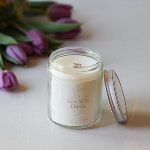 Candle in a glass jar labeled 'Sea Salt & Oasis' with purple tulips in the background.