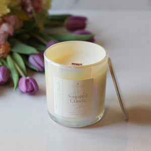 Candle labeled 'Sugared Lemon' with a bouquet of flowers in the background on a light surface.