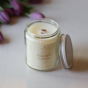 Candle in a glass jar labeled 'Sugar Lemon' with a lid on a light surface, surrounded by purple tulips.