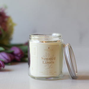 Glass candle jar labeled 'Sugared Limon' with a bouquet of flowers in the background